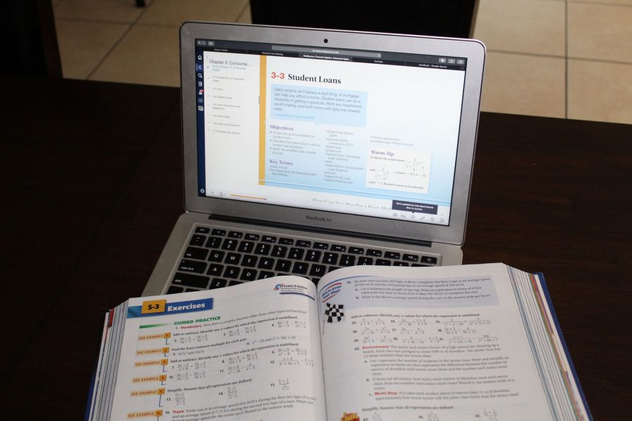 An open textbooks lays on top of a computer which shows an online version of the textbook.