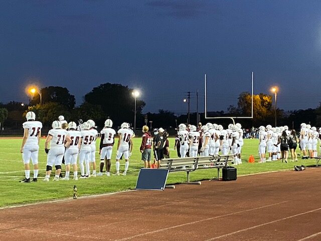 On+their+designated+sideline+of+Colts+Stadium%2C+players+and+coaches+from+the+Stoneman+Douglas+varsity+football+team+stand+shoulder-to-shoulder.