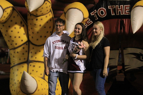 Senior Kacie Shatzkamer stands in between her parents while wearing his MSD Cheer uniform and senior sash. She is holding a bouquet of flowers. The photo was taken in front of the MSD Eagles football teams blow-up entrance.