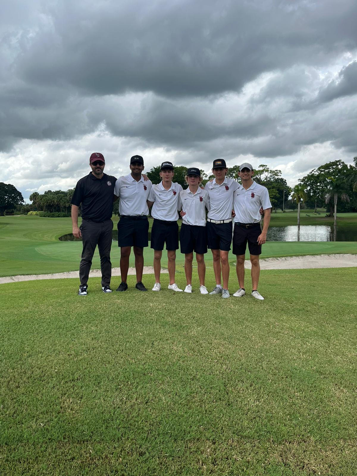 The Marjory Stoneman Douglas mens golf team poses in front of hole nine at Fort Laurdadale Country Club - South Course, following the duration of their round. On the left is Head Coach Darren Levine, followed by senior Jayden Thurasingham, sophomore Josh Lieberman, junior Evan Mazenac, junior Carson Meredith and senior Ryan Shimony.